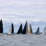 Seven boats, some with black sails, line up for a race